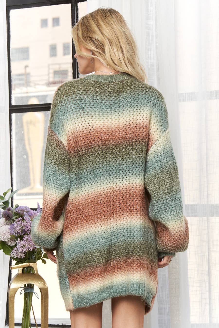 A woman viewed from behind wearing an oversized chunky-knit sweater with horizontal stripes in muted teal, dusty rose, beige, and olive tones, standing indoors near a window.