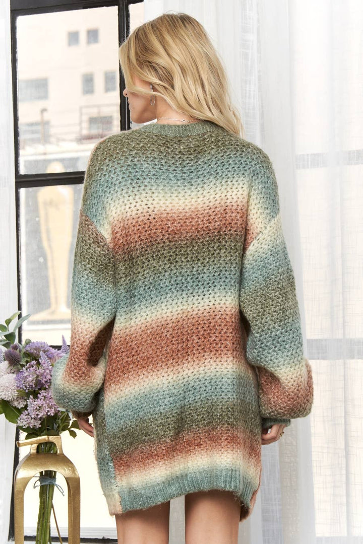 A woman viewed from behind wearing an oversized chunky-knit sweater with horizontal stripes in muted teal, dusty rose, beige, and olive tones, standing indoors near a window.