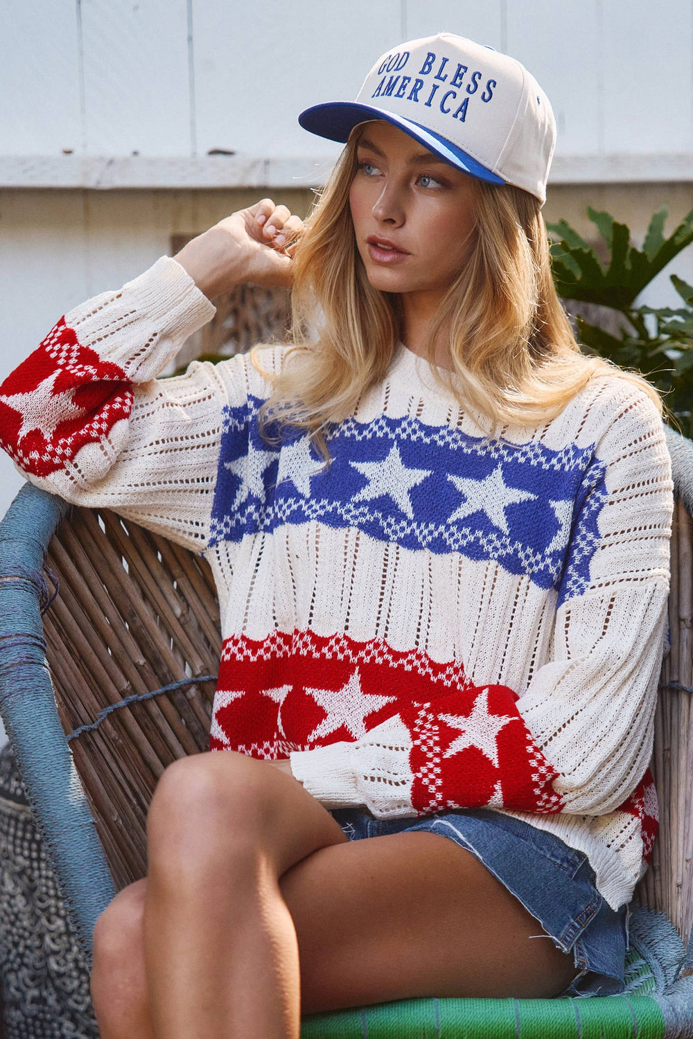Model wearing lightweight ivory open knit sweater with one red stripe and one blue stripe and ivory stars worn with denim shorts 