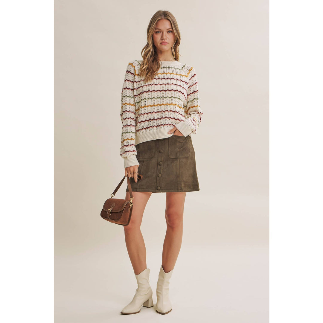 A woman stands against a plain light beige studio background wearing a cream knit sweater with multicolor wavy stripes, a dark olive mini skirt, white ankle boots, and carrying a brown handbag.