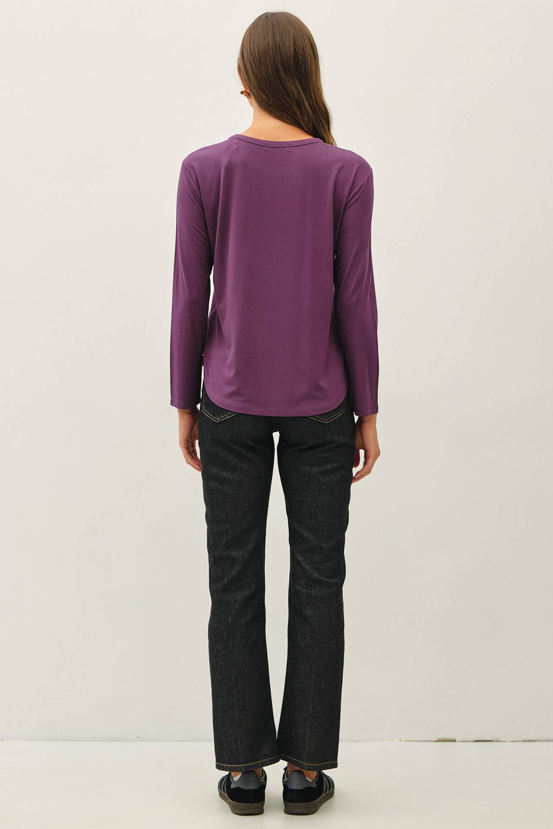 A woman viewed from behind wearing a purple long-sleeve top and dark charcoal gray jeans, standing against a plain off-white studio background.