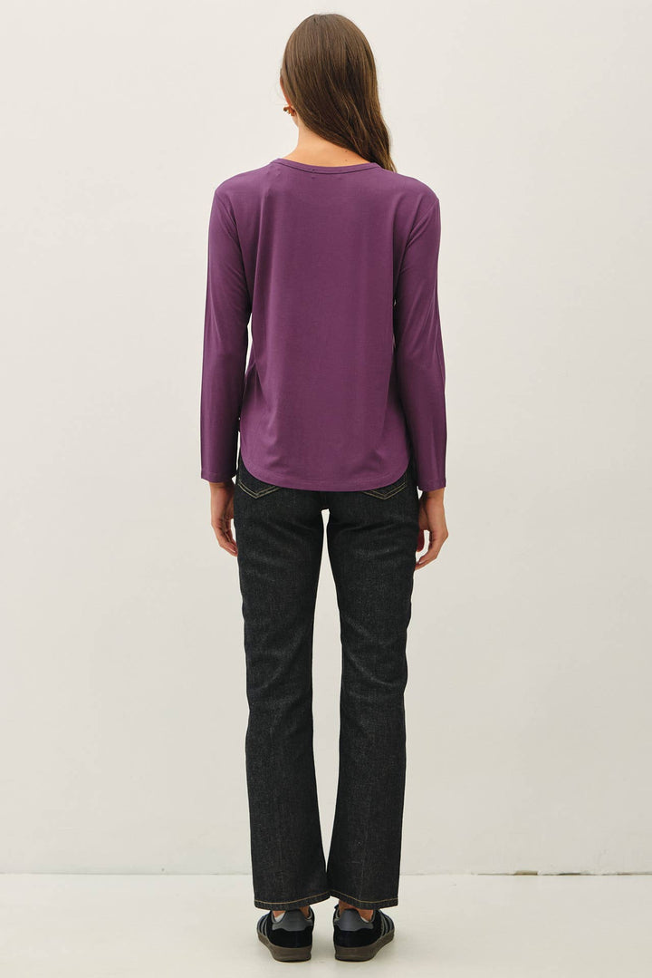 A woman viewed from behind wearing a purple long-sleeve top and dark charcoal gray jeans, standing against a plain off-white studio background.