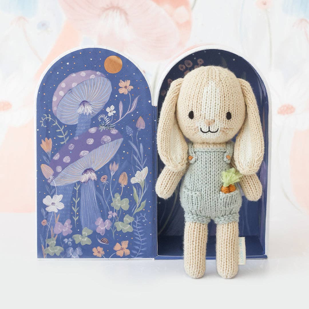 Knitted bunny toy in a green outfit with a decorative box featuring floral and mushroom design.