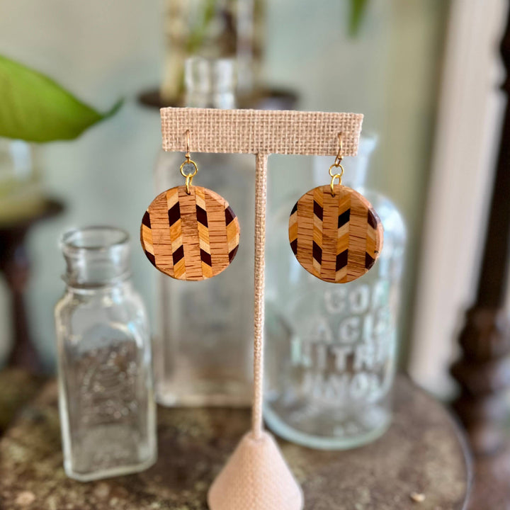Handcrafted wood laminate circle earrings featuring geometric natural wood patterns and hypoallergenic stainless-steel hardware displayed on an earring stand. 
