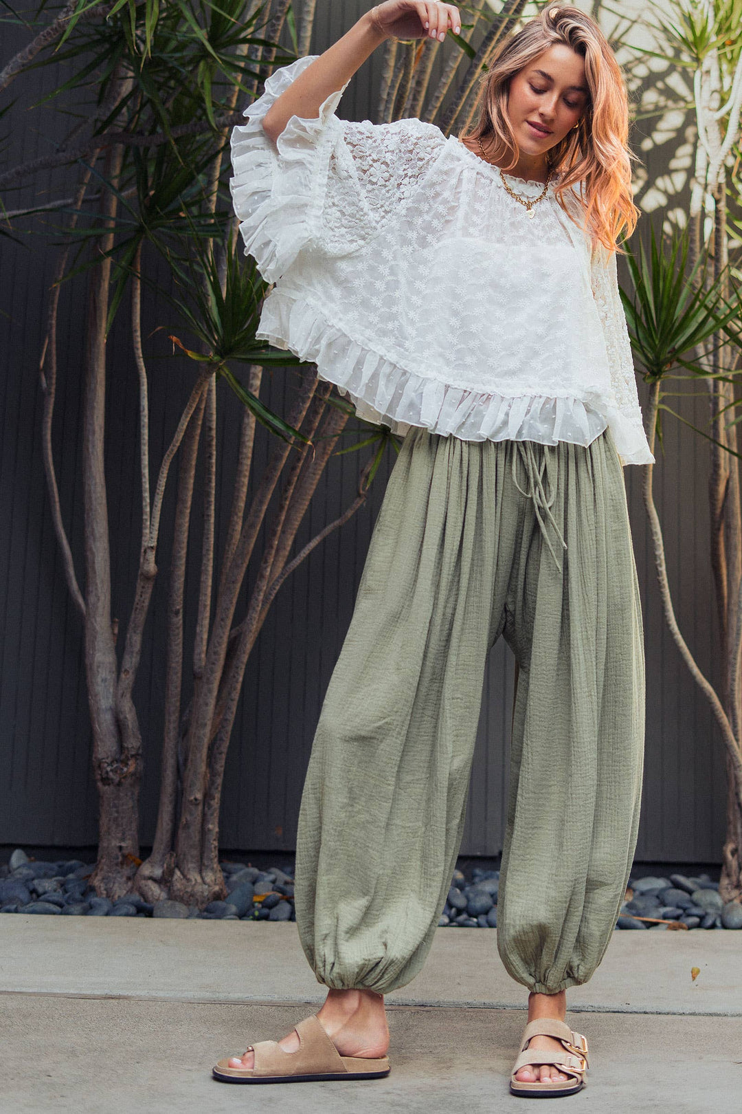 Woman wearing olive green crinkle cotton balloon pants with drawstring waist and elastic ankle cuffs paired with a white lace blouse.