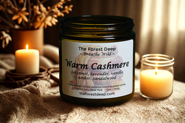 Jar of 'Warm Cashmere' candle with label, surrounded by candles and dried plants on a textured surface.
