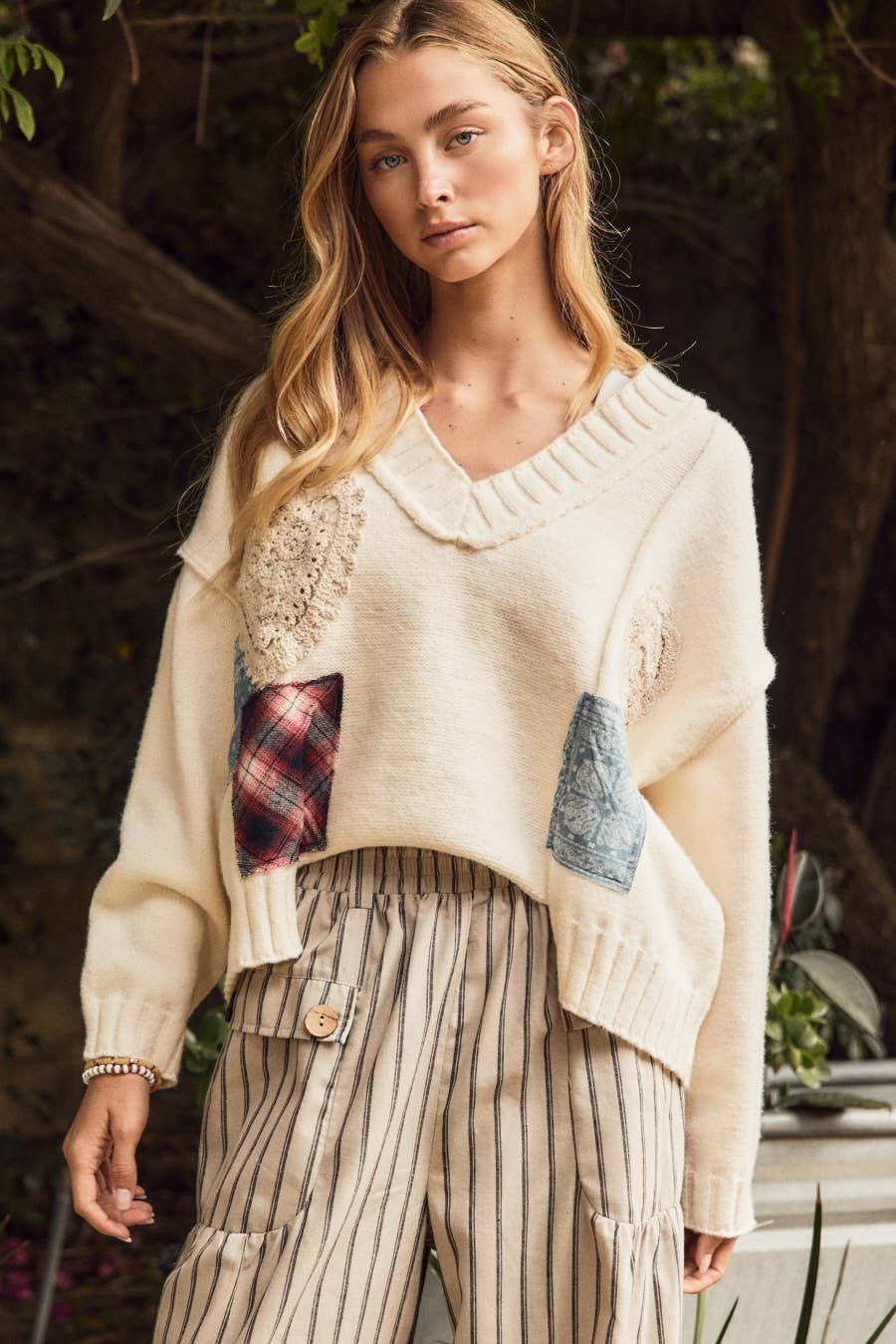 A woman wearing a cream knit sweater with textured lace and patchwork details, paired with beige and brown vertically striped trousers, set against a dark, outdoor foliage background.