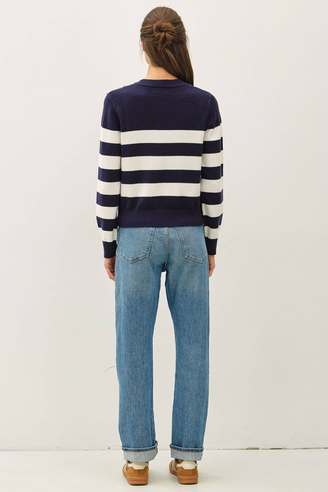 A woman viewed from behind wearing a navy and white horizontally striped long-sleeve sweater, light-wash blue jeans, and brown shoes, standing against a plain off-white studio background.
