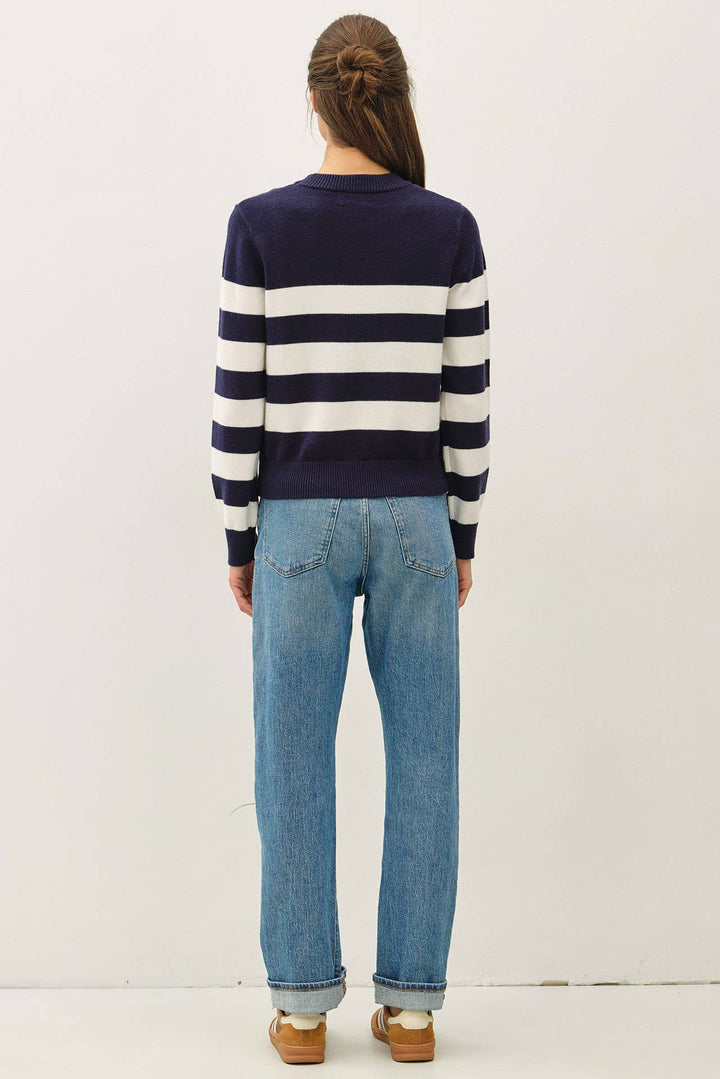A woman viewed from behind wearing a navy and white horizontally striped long-sleeve sweater, light-wash blue jeans, and brown shoes, standing against a plain off-white studio background.