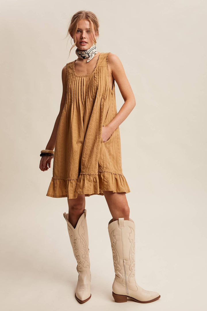 Woman wearing a textured camel-colored sleeveless mini dress with pintuck details and ruffle hem.