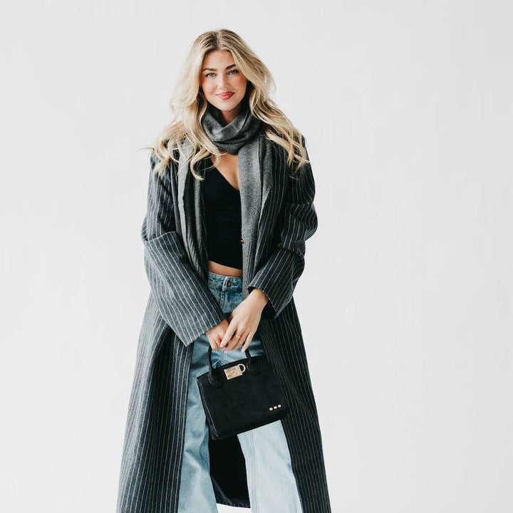 Woman wearing a long gray coat with a black scarf and holding a black handbag on a white background