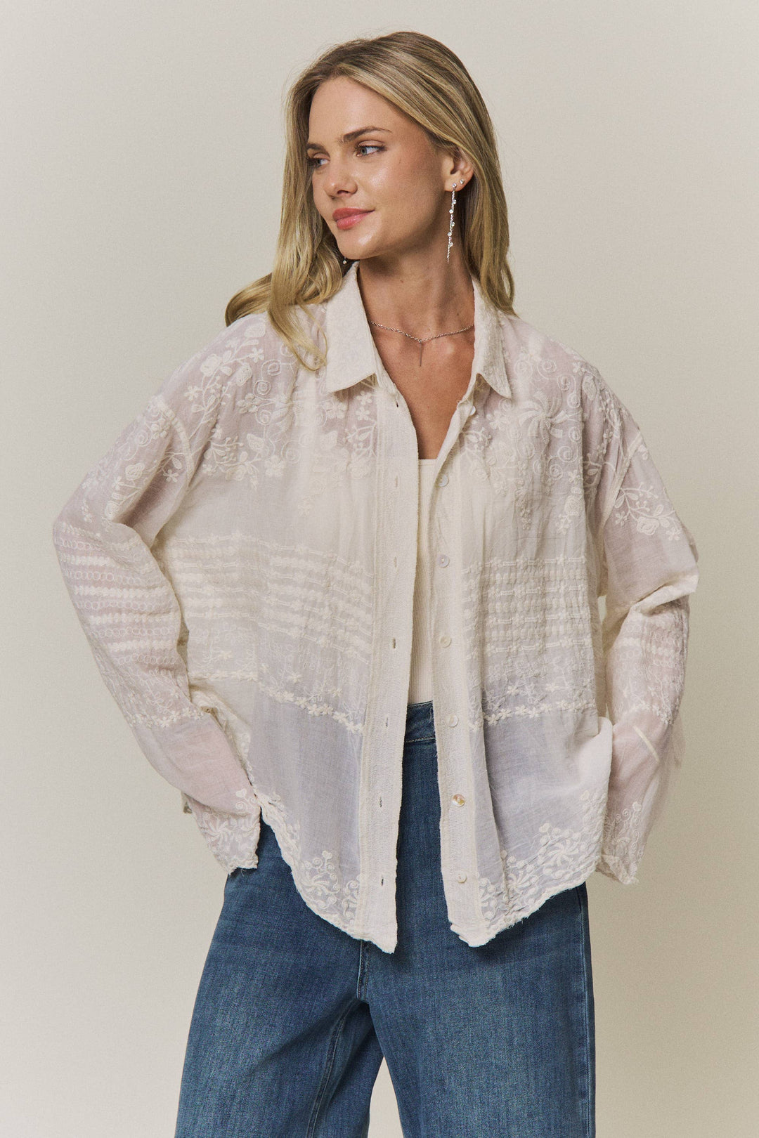 Woman wearing a light-colored lace embroidered blouse and blue jeans against a plain background