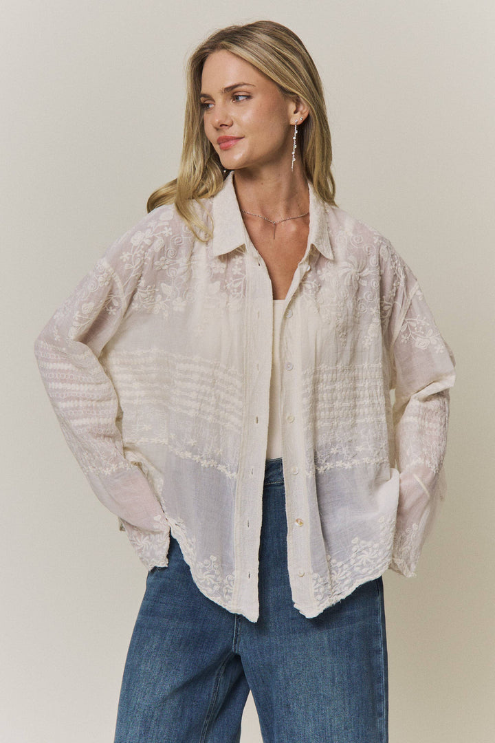 Woman wearing a light-colored lace embroidered blouse and blue jeans against a plain background