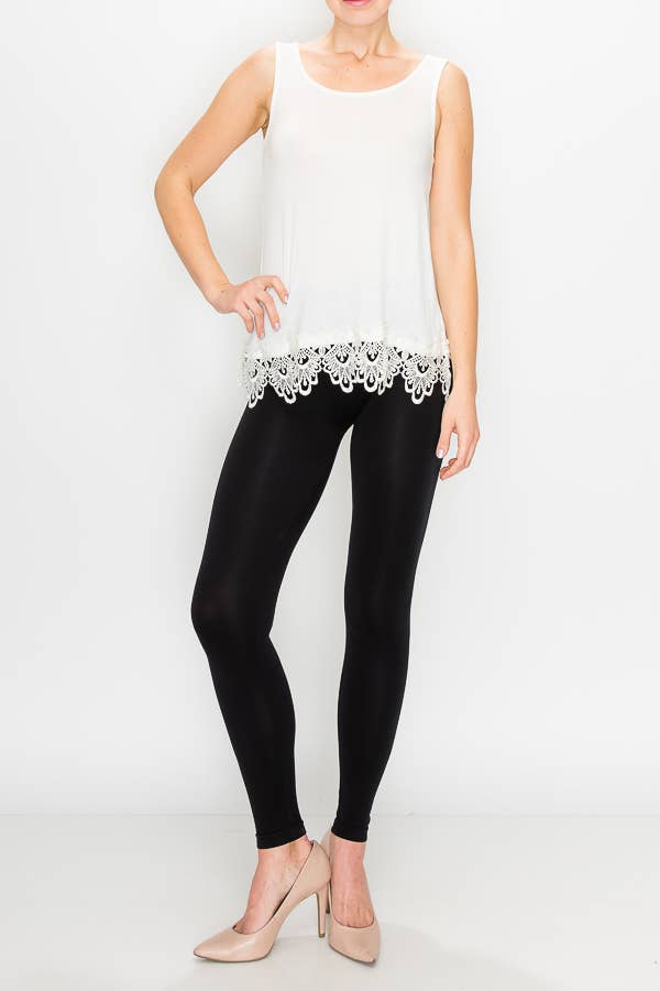 A model wears a sleeveless, scoop-neck tank top in ivory with a wide, intricate lace trim along the bottom hem. She is pairing it with black leggings and neutral-colored heels.

