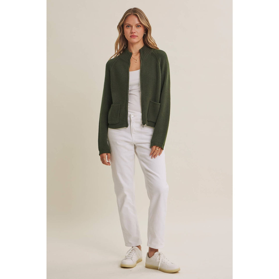 Woman wearing a sweater green jacket, white top, and white pants on a beige background