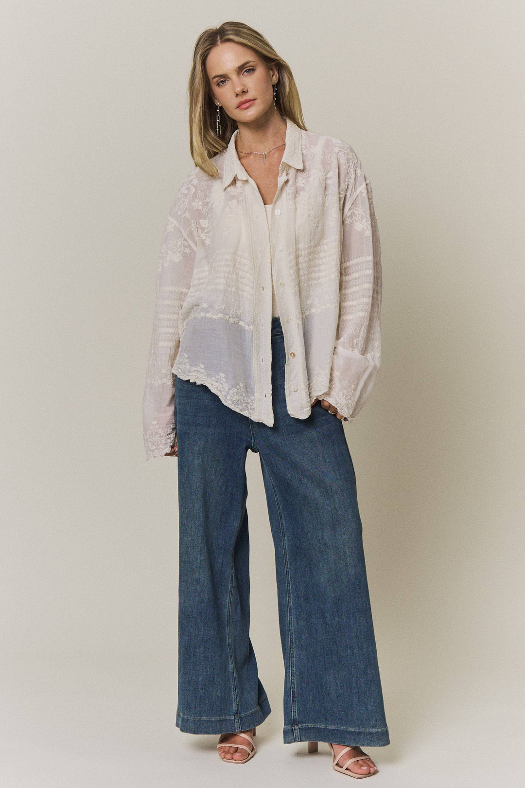 Woman wearing a light-colored lace embroidered blouse and blue jeans against a plain background