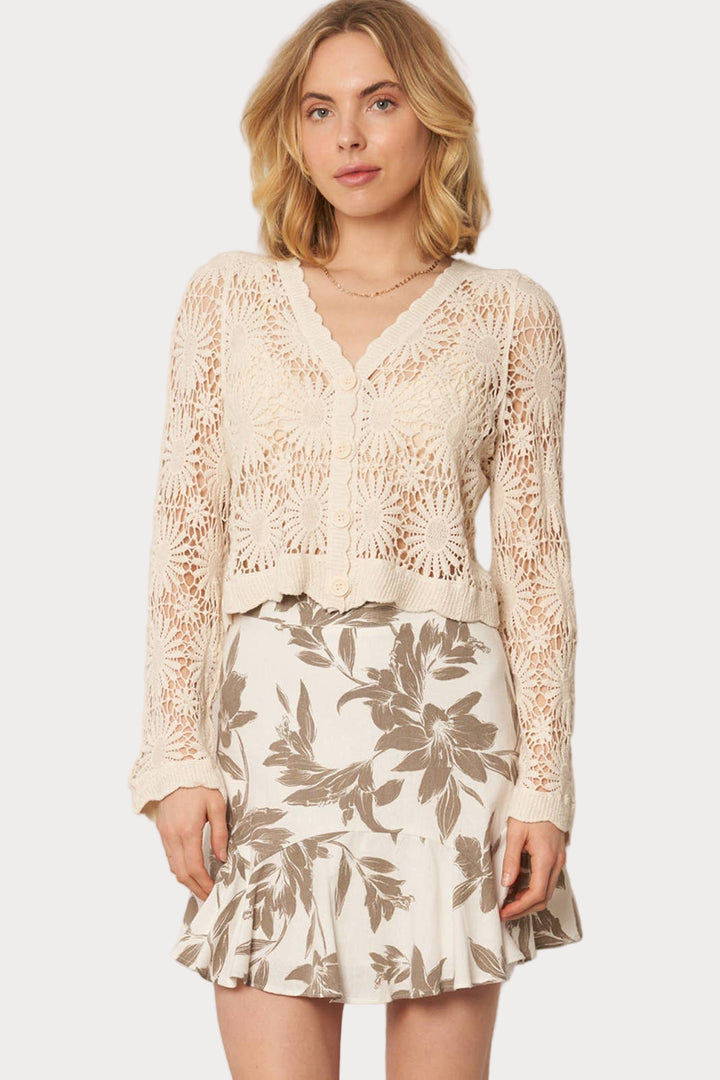 A woman wearing a beige flower pattern crochet cardigan with long sleeves, paired with a white skirt featuring a floral pattern.