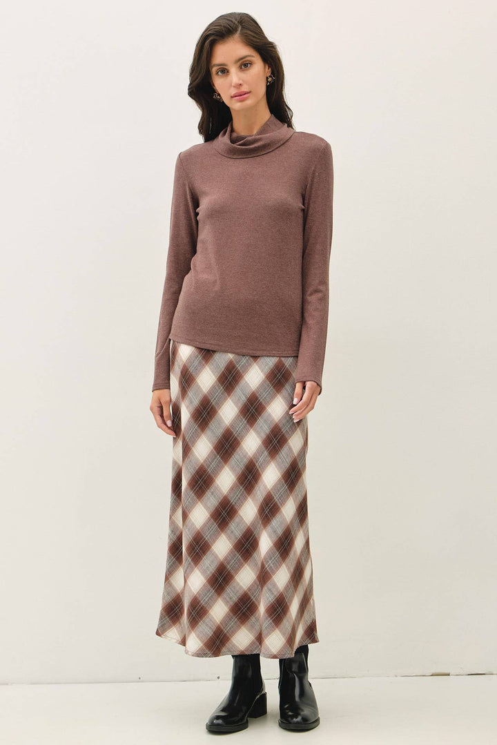 A woman modeling a long-sleeve turtle-neck knit sweater in a dusty mauve-brown color, paired with a brown, beige, and white plaid maxi skirt and black ankle boots, against a plain off-white studio background.