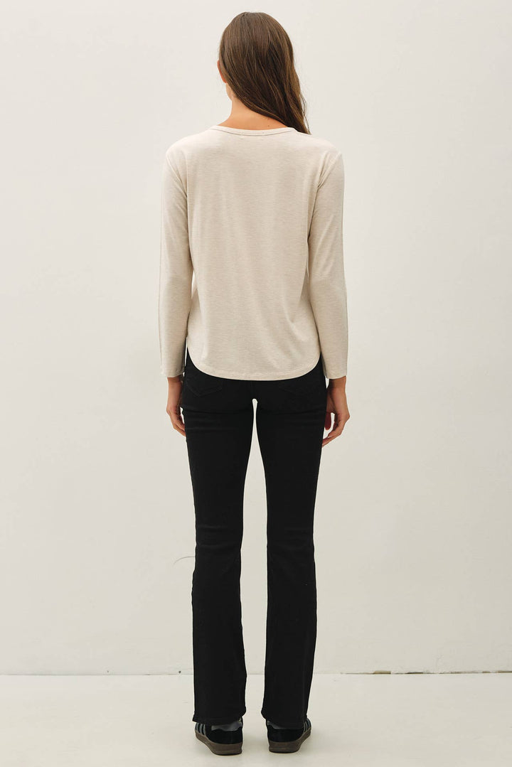 A woman viewed from behind wearing a light cream long-sleeve top and black pants, standing against a plain off-white studio background.