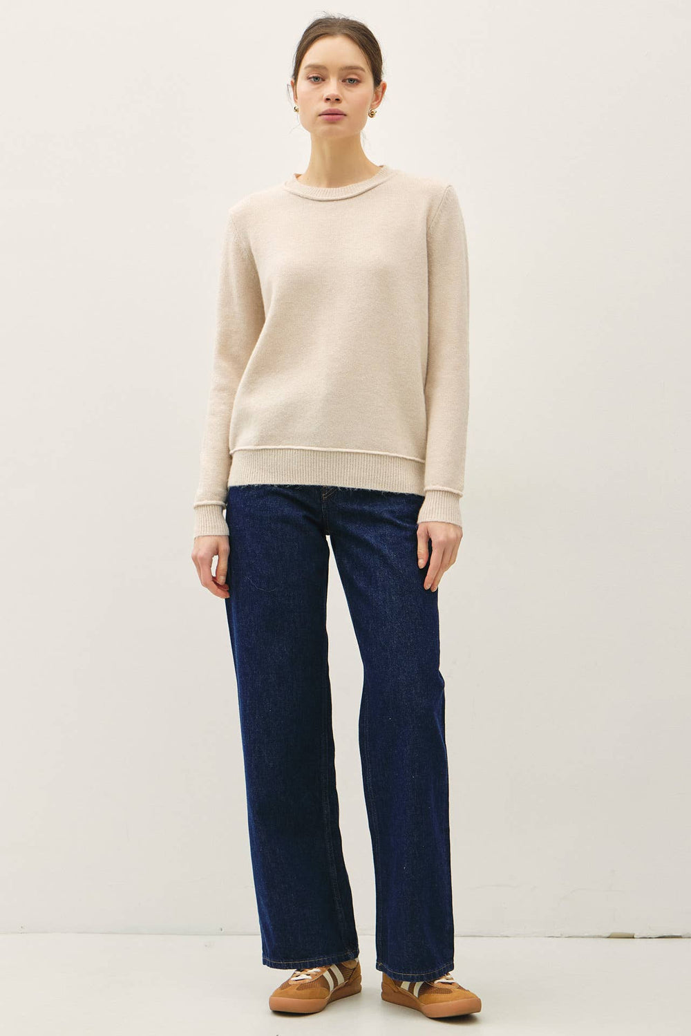 A woman modeling a light beige crewneck sweater and dark blue wide-leg jeans with tan loafers, standing against a plain off-white studio background.