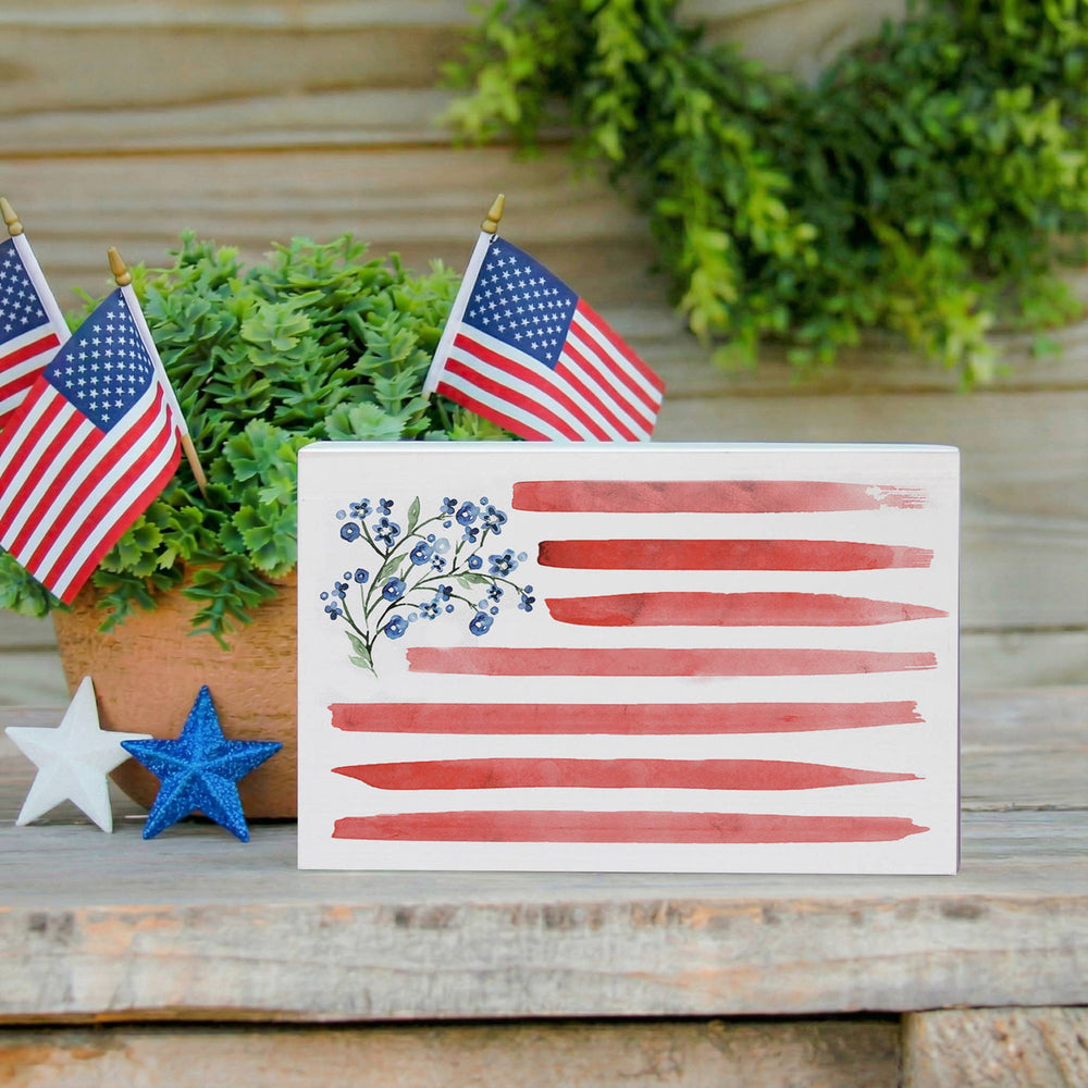Decorative American flag block with small American flags and greenery on a wooden surface.