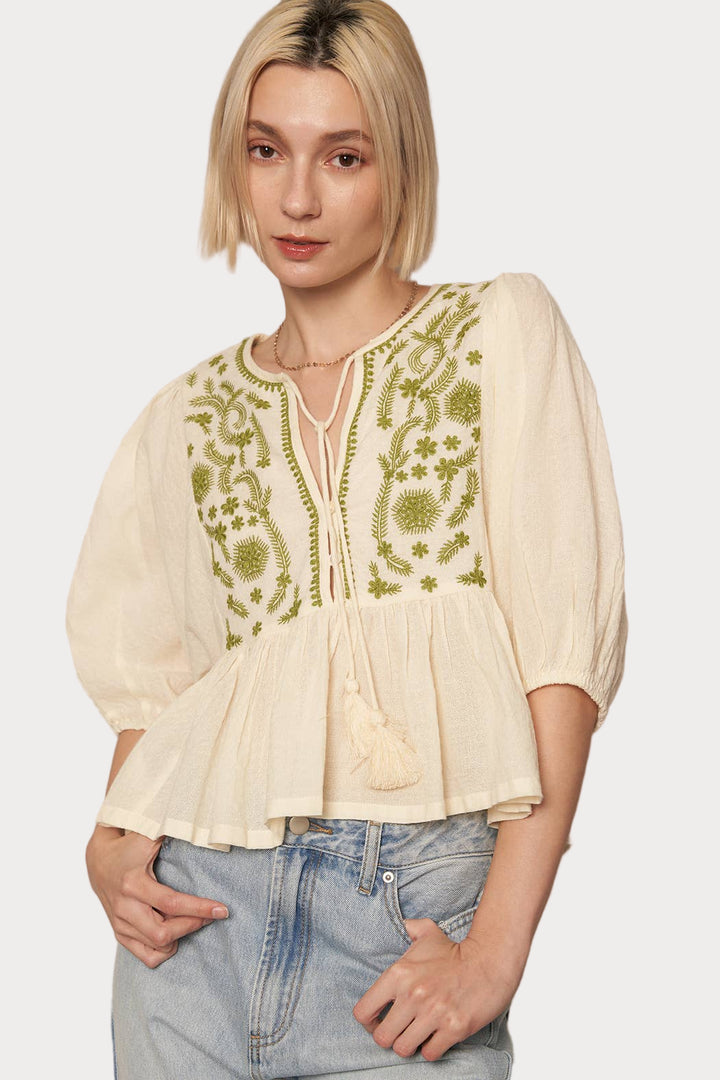 A woman wearing a beige peplum top with puff sleeves and green embroidery details, paired with blue jeans.