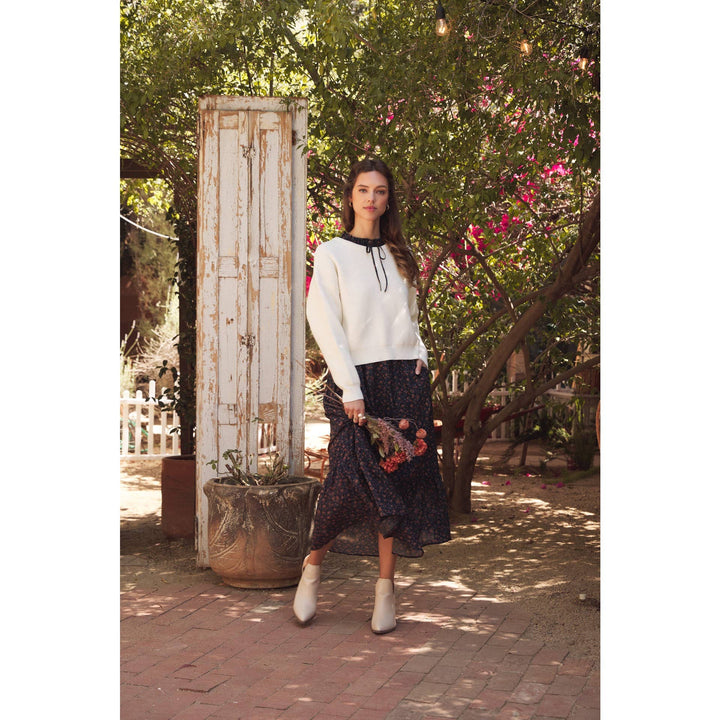 Model wearing Bow Back Sweater | Dolman Sleeve Knit with Satin Tie. Lifestyle photo with outdoor garden setting.  Distance shot showing sweater layered with a black blouse paired with floral skirt & neutral booties.