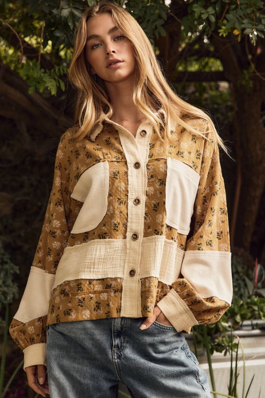 A woman wearing a patchwork button-up shirt with a brown floral print and cream-colored panels, paired with blue jeans, set against a lush green outdoor background.