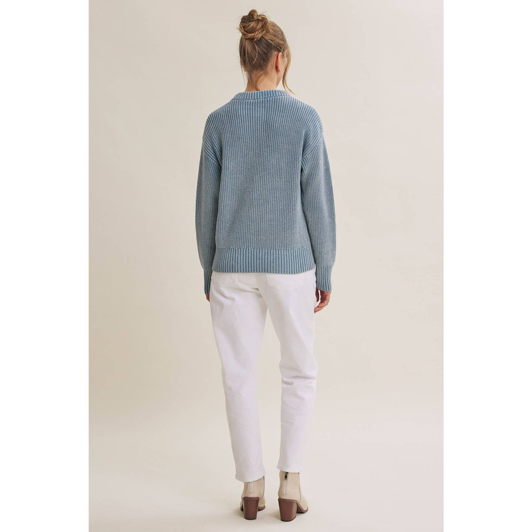 A woman viewed from behind wearing a light blue ribbed knit sweater, white pants, and beige heels against a plain beige studio background.