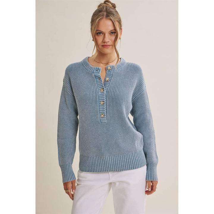 A woman wearing a light blue ribbed knit sweater with a buttoned henley neckline and white pants, standing against a plain beige studio background.