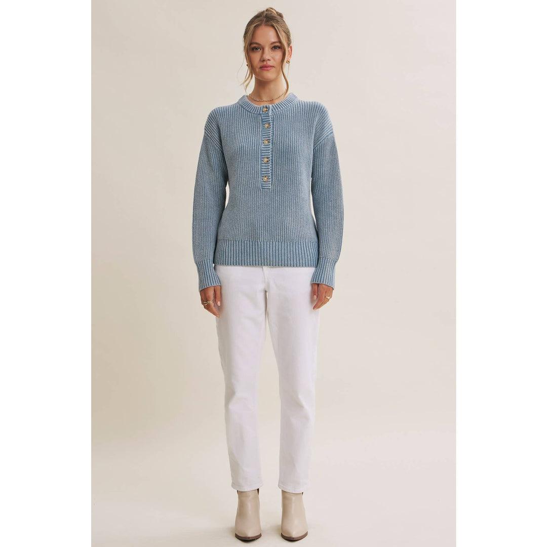 A woman modeling a light blue ribbed knit sweater with a buttoned henley neckline, paired with white pants and beige ankle boots, against a plain beige studio background.