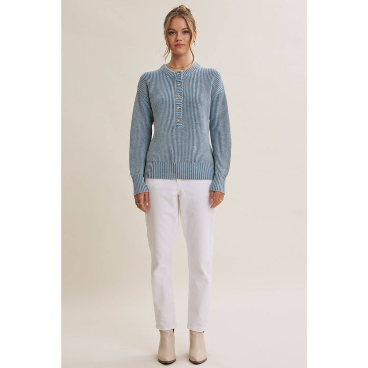 A woman modeling a light blue ribbed knit sweater with a buttoned henley neckline, paired with white pants and beige ankle boots, against a plain beige studio background.
