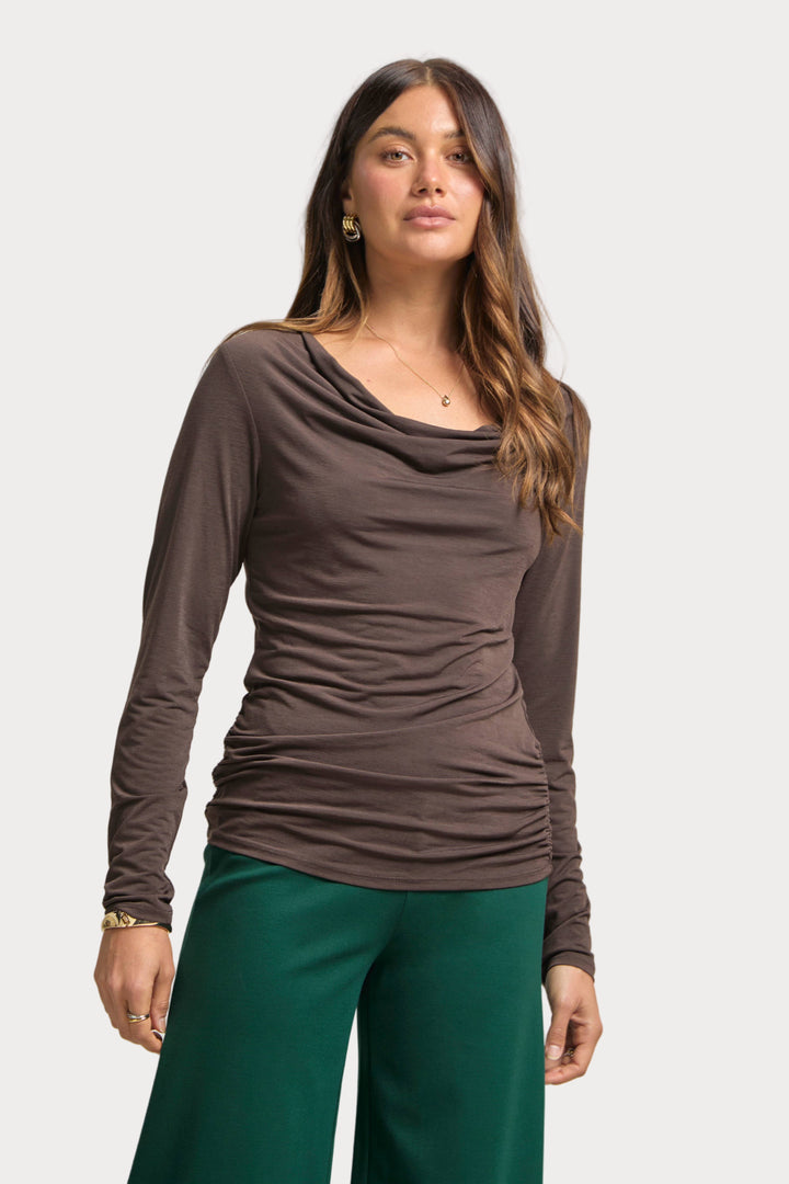 Woman wearing a brown long-sleeve top and green pants on a plain background
