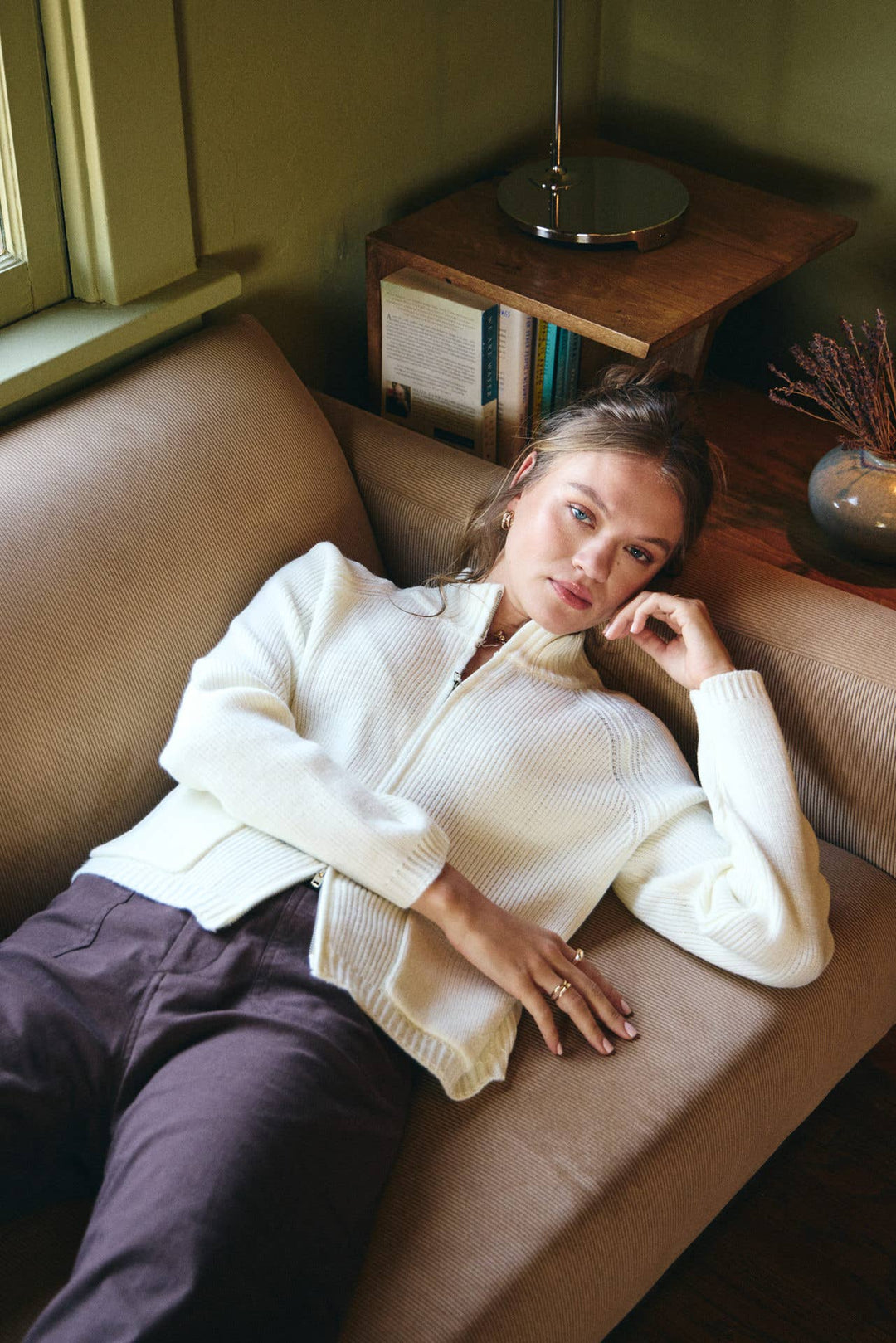 Woman in a white sweater and dark pants lying on a brown couch in a cozy room.