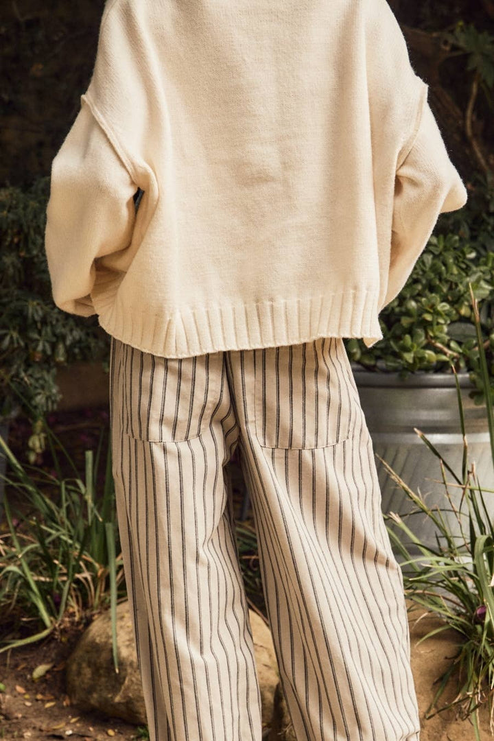 A person outdoors wearing a cream knit sweater and beige wide-leg trousers with thin dark vertical stripes.