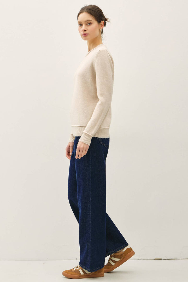 A woman modeling a cream long-sleeve knit sweater and dark blue wide-leg jeans, paired with brown leather shoes, against a plain off-white studio background.