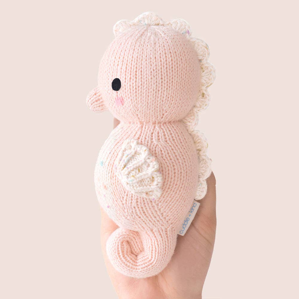 Knitted pink seahorse toy held by a hand against a light background