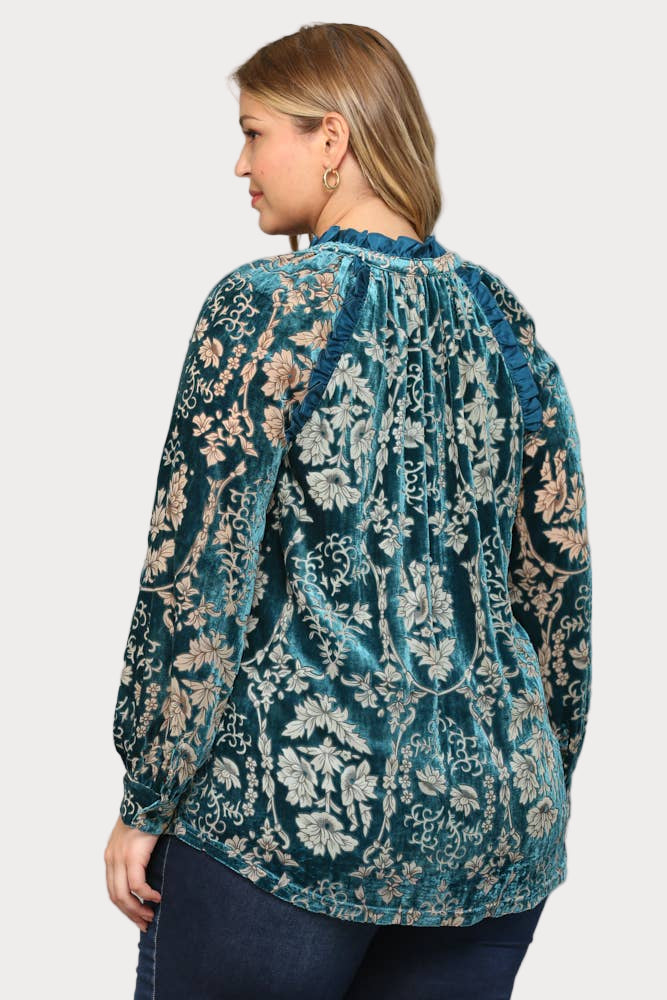 A smiling plus-size model wears a long-sleeve teal burnout velvet blouse featuring a floral pattern, ruffle trim, and a tassel tie at the neck.

