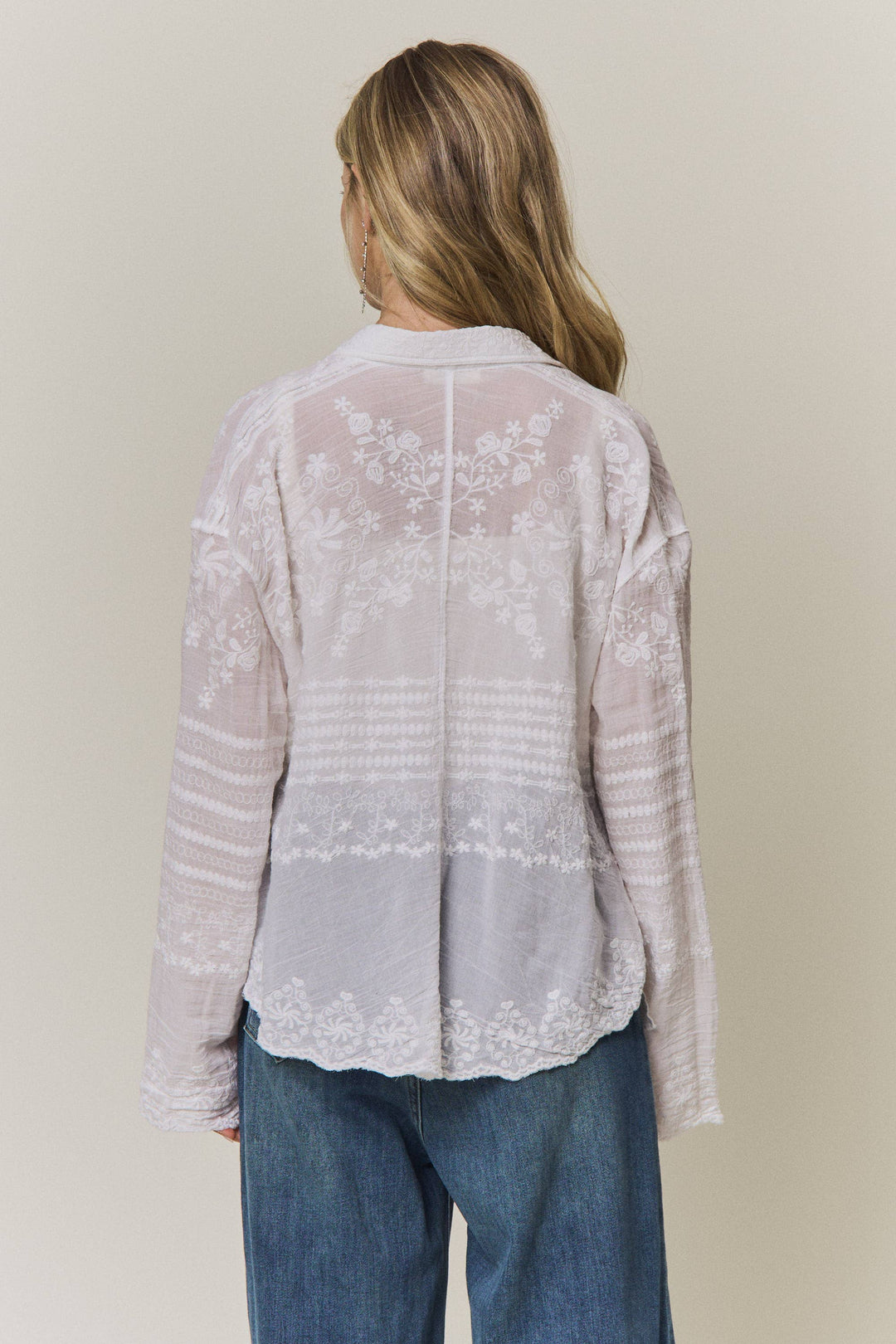 Woman wearing a light-colored lace embroidered blouse and blue jeans against a plain background