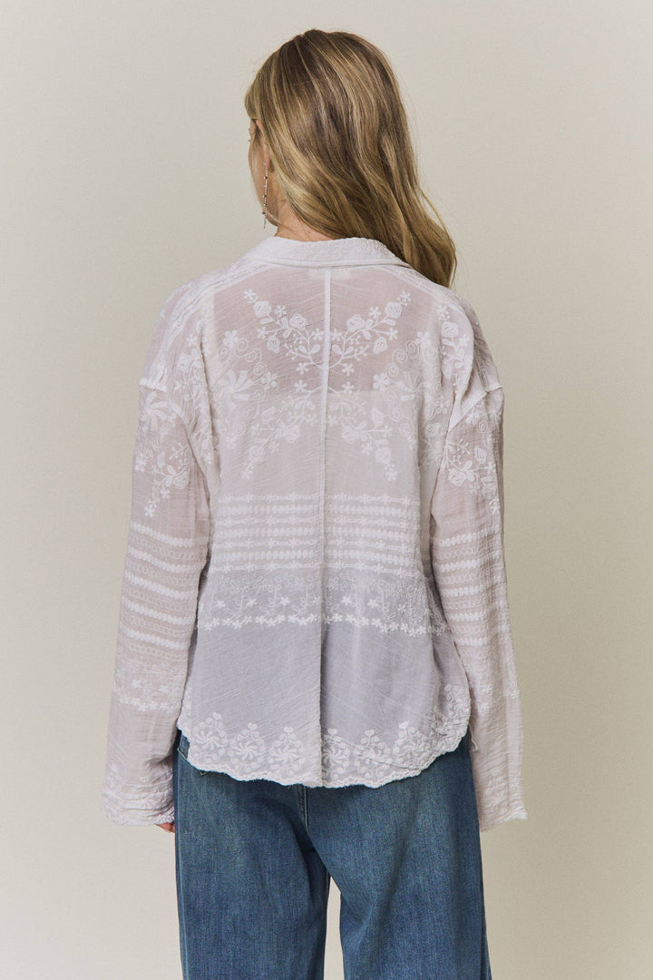 Woman wearing a light-colored lace embroidered blouse and blue jeans against a plain background