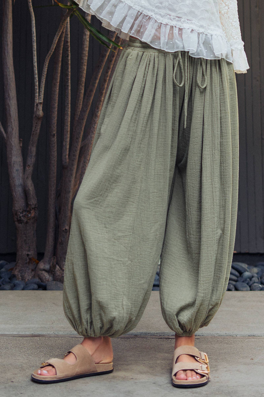 Woman wearing olive green crinkle cotton balloon pants with drawstring waist and elastic ankle cuffs paired with a white lace blouse.