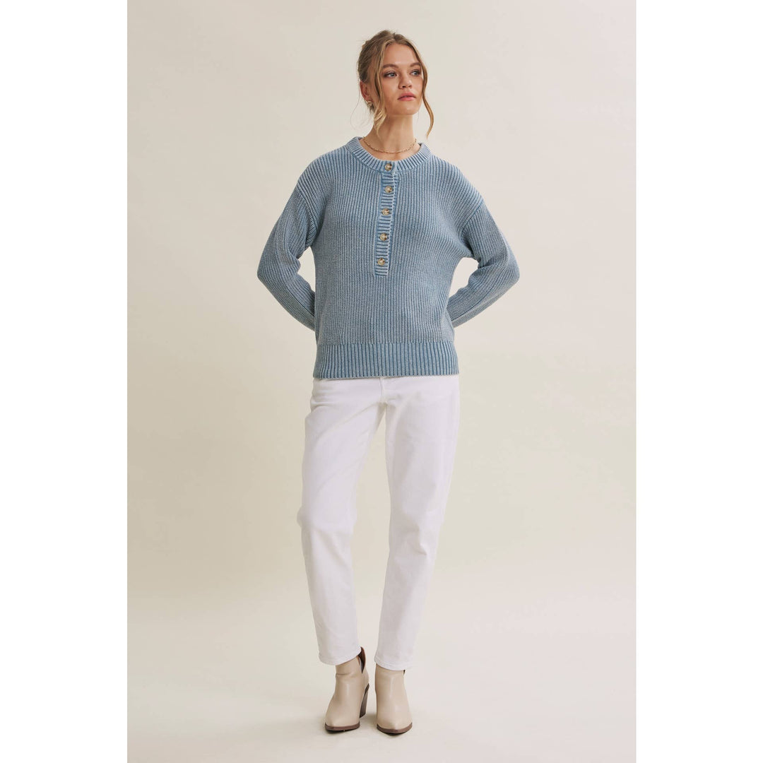 A woman modeling a light blue ribbed knit sweater with a buttoned henley-style neckline, paired with white pants and beige ankle boots, against a plain light beige studio background.