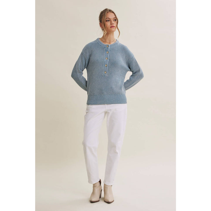 A woman modeling a light blue ribbed knit sweater with a buttoned henley-style neckline, paired with white pants and beige ankle boots, against a plain light beige studio background.