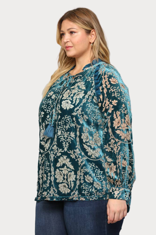 A smiling plus-size model wears a long-sleeve teal burnout velvet blouse featuring a floral pattern, ruffle trim, and a tassel tie at the neck.

