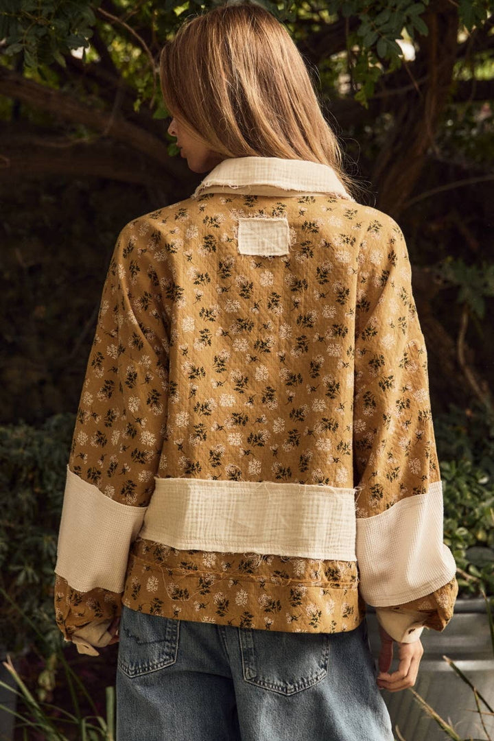 A woman viewed from behind wearing a tan and brown floral patterned jacket with contrasting cream-colored collar, cuffs, and a horizontal band, paired with blue jeans in an outdoor garden setting.