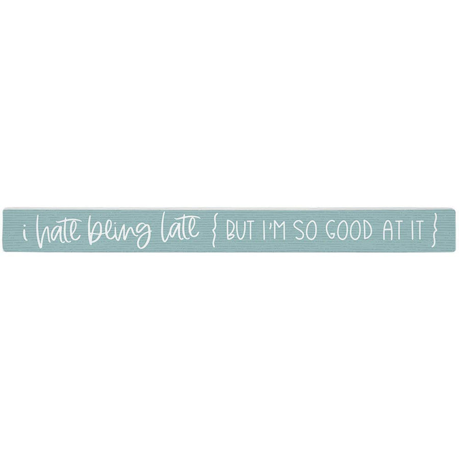 Textual phrase 'i hate being late { BUT I'M SO GOOD AT IT }' on a light blue wooden block. Shown on a white background