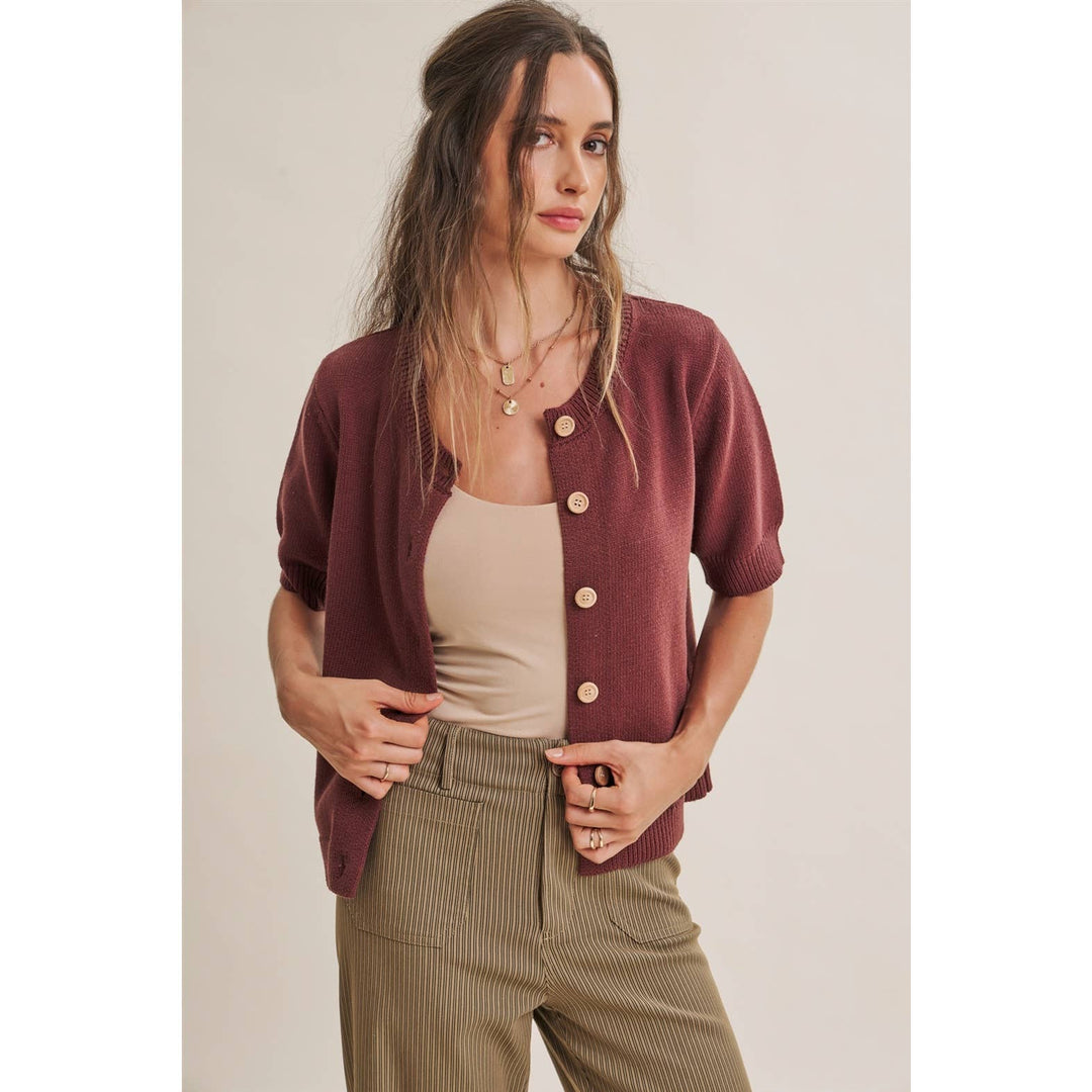A woman modeling a short-sleeve button-front cardigan in burgundy over a beige tank top and brown stripped trousers, photographed against a plain light beige studio background.