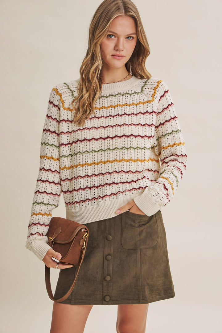 A woman modeling a cream knit sweater with horizontal stripes in burgundy, mustard yellow, and olive green, paired with an olive green button-front skirt and holding a brown leather crossbody bag against a warm beige studio background.