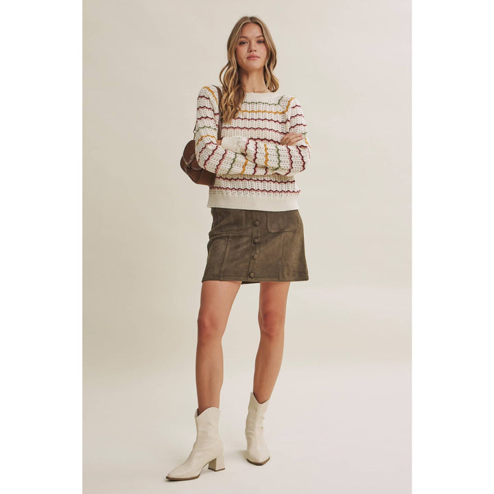 A woman modeling a cream knit sweater with multicolor horizontal stripes, a brown mini skirt, and off-white ankle boots, standing against a solid light beige studio background.