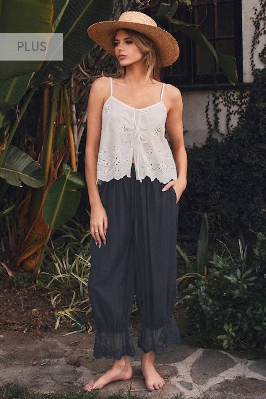 Woman wearing black relaxed lace hem pants with an elastic drawstring waistband and scalloped lace cuffs paired with a white embroidered camisole.